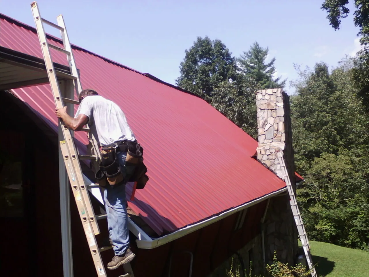 Roofer on a ladder beside a red metal roof and stone chimney — Soffit & Fascia Repair by Roofing Brothers in Scott